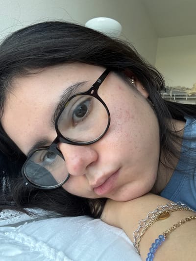 A close-up, upside-down portrait features a person with dark hair and fair skin, likely indoors in Austin, United States of America. The subject is wearing round, tortoise-shell eyeglasses, through which one eye with dark blue or grey iris and black eyeliner is clearly visible. The other eye is partially obscured by the glasses frame. The skin shows texture with visible pores and some minor blemishes on the forehead and cheek.
The person is resting their head on their hand or arm, conveying a relaxed posture. They wear multiple pieces of jewelry: small, shiny earlobe piercings (one appearing as a silver ball stud, another as a small gold hoop), and layered necklaces. The necklaces include a fine gold chain, a silver chain with a small, circular golden charm, and a beaded necklace with light blue, faceted stones.
The subject is wearing a light blue top, possibly ribbed, and a portion of a white garment with subtle texture is visible on the right side of the frame. The background is softly blurred, indicating an interior setting, possibly with ambient natural light. The brand "Ray-Ban" is faintly visible on the arm of the eyeglasses. No specific activity beyond resting is depicted.