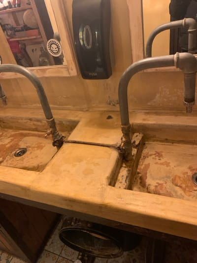 An interior shot, oriented sideways, captures a rustic utility or washroom area featuring two integrated basins and exposed plumbing. The walls and basins are constructed from a material with a textured, worn, and earthy appearance, possibly concrete or plaster, showing signs of aging or distressed paintwork.
The primary subjects are the grey-colored, exposed metal pipes, likely galvanized steel, which form intricate U-bends and connect to brass fittings. One set of pipes extends upwards from the left, while another set feeds into the lower basin area. A thinner, darker pipe runs vertically, connecting the plumbing systems of both basins.
On the left wall, a black, wall-mounted soap dispenser is visible, bearing the word "PUSH" in white text. Below it, a mirror reflects shelves holding various objects and a white circular sticker featuring a stylized, smiling face. To the far right of the image, parts of a dark, round container or fixture are visible next to a wall adorned with patterned tiles, suggesting a more decorative element beyond the immediate sink area.
The overall atmosphere is dim, indicating an indoor setting with subdued lighting. No activity is taking place, and there are no people present. The scene conveys a functional yet aged and industrial aesthetic. The specific location provided, Marousi, Greece, helps contextualize the style.