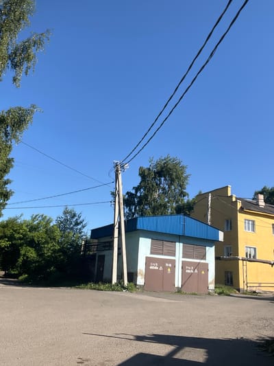 A low-angle, vertical, eye-level shot shows a street with a concrete power pole and overhead wires in the foreground. Behind the pole is a small, light blue building with a blue, corrugated metal roof and two large brown garage doors. The garage doors have warning symbols and text. To the right of the blue building is a tall, yellow apartment building with several windows. Trees and foliage are visible in the background and on the left side of the image. The sky is clear and blue. The scene is likely in Kiselyovsk, Russia, based on similar imagery.