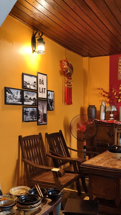 This is a medium-shot, eye-level, indoor photograph capturing a corner of a restaurant or cafe, likely in Lam Vien - Da Lat, Vietnam, based on the framed text and images. The walls are painted a warm mustard yellow, and the ceiling is paneled with dark wood.
A vintage-style wall lamp with a metal base and glass shade emits a warm light on the left side of the wall. Below the lamp, a collage of framed black and white photographs is arranged. One of the frames contains the text "DA LAT" in bold capital letters. A decorative red lantern with gold accents and red tassels hangs from the ceiling near the wall.
In the foreground, two dark wooden chairs with slatted backs face inwards towards a wooden table. On the table, there are several dark bowls and plates, some with remnants of food and chopsticks. A close-up of the bowls shows they are black with some intricate woven detailing on the stands.
To the right of the chairs, a tall black floor fan with a red center is positioned behind the chairs. Behind the fan, a dark wooden cabinet or dresser is visible. On top of the cabinet, there are several objects, including a dark ceramic vase, a silver container, and a small lamp with a decorative shade. A branch with red flowers and green leaves is placed behind these items, adding a splash of color.
The overall atmosphere suggests a cozy, retro-themed establishment. The lighting is warm and inviting, and the vintage decor contributes to a sense of nostalgia. The focus is on the seating area and the decorative elements on the walls and cabinet, with the table in the foreground showing signs of recent use.