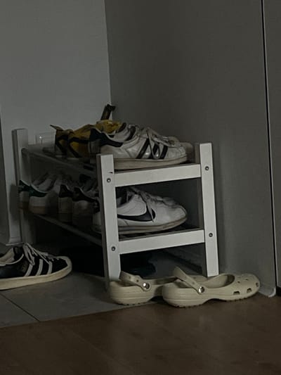 This media file shows a close-up, rotated view of shoes on a multi-tiered white shoe rack. The rack is positioned against a dark gray, smooth surface, possibly a wall or cabinet, with a thin horizontal line visible near the top, suggesting a seam or edge. To the right of the shoe rack, a brown, textured surface is visible, which might be a wooden floor or wall.

On the shoe rack, several pairs of footwear are displayed. On the top tier, a pair of white sneakers with black stripes and accents are visible. Next to them, another pair of white sneakers with a black Nike swoosh logo is present. On the right side, a pair of light beige Crocs are stacked, one above the other, with the strap down. Below the top tier, more shoes are visible, including a pair of black and white sneakers with stripes on the bottom tier, and another pair of partially visible shoes with yellow and white accents on the second tier from the top.

The lighting in the image is dim, suggesting an indoor setting, possibly a closet or entryway, at an unspecified time of day. There is no visible text in the image. The scene depicts an organized storage of footwear.