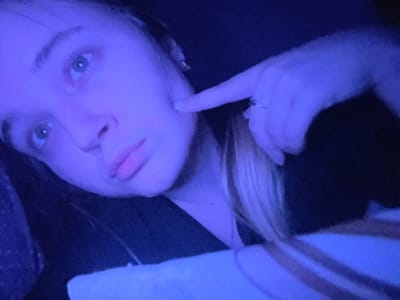 The image presents a close-up of a female subject under strong blue ambient lighting, resulting in a monochromatic blue cast over the scene. The subject's head and upper torso are visible, positioned at an upward angle, with her gaze directed towards the upper left. Her right index finger, adorned with a ring featuring multiple small stones, is lightly pressed against her right cheekbone. The subject has light blonde hair, visible over her right shoulder and partially framing her face, and light-colored eyes. She appears to be wearing dark clothing. In the lower right corner, a light-colored textile object, possibly a blanket with faint stripes, is present. The background is uniformly dark and indistinct, offering no specific environmental details. The image contains no visual information to confirm the specified location as Pushchino, Russia.