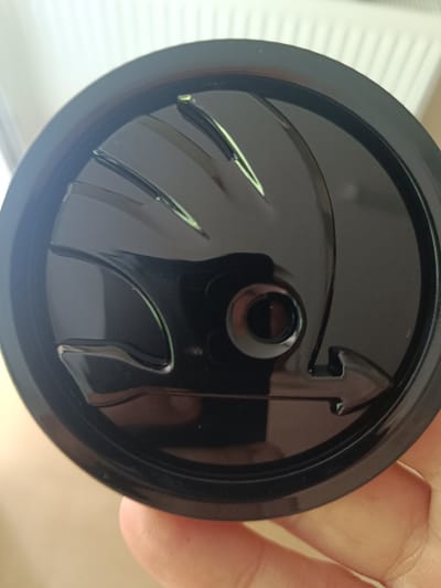 A black, circular car emblem with a wing motif is being held by a person's hand. The emblem is shiny and reflects light, showing some texture and raised elements. The background is blurred but appears to be indoors with a white, ribbed surface, possibly a radiator or air vent. The image does not contain any information about the city of Huntingdonshire, United Kingdom.