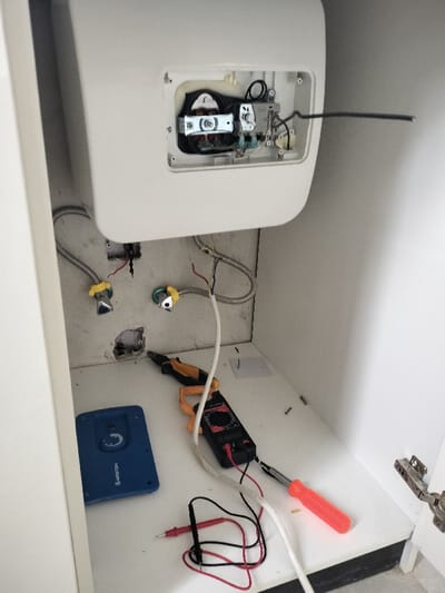 The scene depicts the interior of a white utility cabinet in Falomo, Nigeria, indicating ongoing or recently completed maintenance. A white wall-mounted appliance, likely an instant water heater, has its front panel removed, exposing internal electrical components, wiring, and a heating element. A black electrical cable extends horizontally from the appliance's right side.

Below the appliance, two flexible corrugated metal water hoses, secured with yellow plastic clamps, connect to wall-mounted valves. Beneath these connections, the wall shows exposed plaster and dark discoloration, consistent with burn marks or charring, surrounding a partially revealed electrical conduit or outlet.

On the white floor of the cabinet, several tools and objects are systematically arranged:
*   A pair of yellow and black combination pliers.
*   A black digital multimeter or clamp meter with a yellow clamp, featuring red and black test leads connected to probes.
*   A red-handled screwdriver with a black tip.
*   A blue rectangular device labeled "ARISTON," possibly an electronic controller or component for the appliance.

Additionally, loose screws and a small piece of white paper are visible on the cabinet floor. The right side of the image partially shows a silver cabinet hinge. No individuals are present in the image.