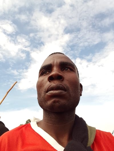 The image shows a young African man, likely in his early 20s, looking upwards towards a partly cloudy sky. He is wearing a red jersey, a white undershirt, and a dark green and black shawl. The man appears to be standing in an open area with a stick lying on the ground in the foreground. This image was taken in Burundu, Kenya, which is a rural village located in the western part of the country, near Lake Victoria, known for its scenic views and natural beauty. The coordinates provided place the image within the village's boundaries. The man's expression is serene and contemplative, suggesting he is possibly lost in thought or reflecting on the scenery. There are no other people or objects in the frame, suggesting he is alone at this moment.