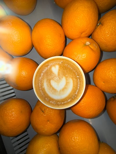 An overhead shot presents a white cup of coffee, likely a latte or cappuccino, prominently centered in the frame. The coffee's surface features intricate latte art, resembling a stylized heart or leaf design within its creamy foam.
Surrounding the coffee cup, a multitude of vibrant orange oranges fills the remainder of the image. These oranges appear fresh and whole, some exhibiting minor imperfections or parts of their stems, suggesting a natural and unprocessed look. They are arranged densely and somewhat randomly on a grey surface. In the lower left and right corners of the image, the grey surface shows distinct parallel grates or slats, indicating it might be a cooling rack, a specific type of counter, or a display shelf.
The lighting in the image is bright, creating a soft illumination. A significant bright flare or reflection is visible in the bottom-center area, possibly from a light source. No people are present, and no specific activity is taking place; the image is a static composition of food and drink items. There is no discernible text within the image. This scene is located in Veshki, Russia.