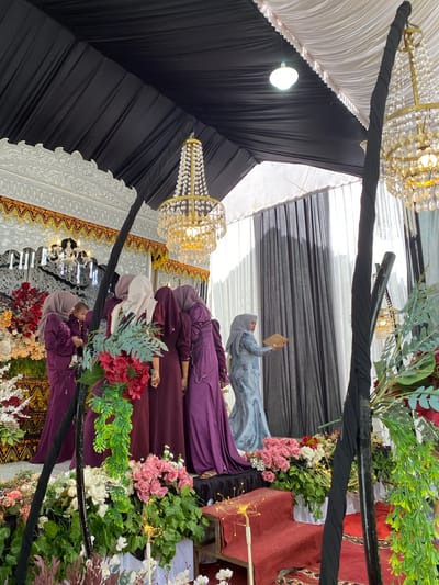 This image captures an indoor event within a temporary structure, likely a tent or pavilion, in Matangbaroh, Indonesia. The scene centers on a raised stage area decorated for an occasion.

The ceiling of the structure is draped with black and white fabric panels, featuring several light fixtures, including two large ornate golden chandeliers with crystal elements. The stage backdrop is elaborate, constructed from white panels with intricate carvings and accented with gold and black patterned borders.

On the stage, approximately six women are present, all wearing traditional long dresses (baju kurung) and hijabs. Most women are dressed in various shades of maroon or deep purple. One woman on the far left holds a young child, who is also dressed in maroon. Another woman, positioned further right, wears a light blue patterned dress and a matching hijab, and is holding a light-colored, book-like object in her hands. The women are standing on the stage, some facing each other.

The stage itself features steps covered with red carpet. The front of the stage and the lower ground area are extensively decorated with floral arrangements, primarily consisting of pink and white roses, mixed flowers, and various green foliage, including hanging vines. Small, warm-toned fairy lights are interspersed within these floral displays. Two prominent black-wrapped vertical poles, likely structural supports, are visible in the foreground and mid-ground, also adorned with foliage. Draped black and white curtains are visible as a backdrop further into the scene on the right.