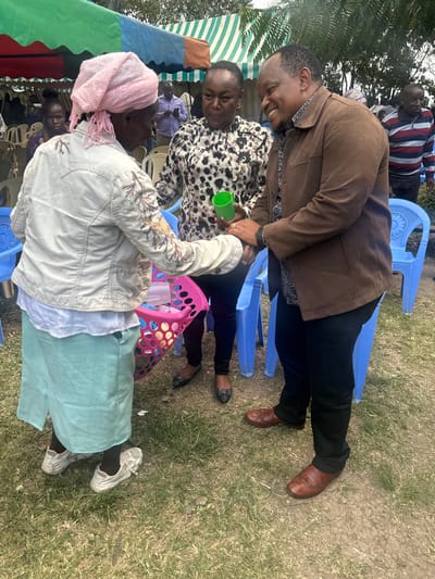 This media file depicts an outdoor social gathering in Kiganjo, Kenya, under bright daylight. The scene is set on a grassy area, furnished with several blue plastic chairs. A striped tent, offering shade, is partially visible on the left side of the frame.

Several adults are present and engaged in interaction. In the foreground, a woman is seen from behind, wearing a light pink head covering, a light-colored jacket with embroidery on the sleeves, a light blue/green skirt, and white shoes. Adjacent to her, a woman with short hair and a leopard print top sits, holding a green plastic cup and looking towards the camera with a subtle smile. Standing between these two, a man in a brown jacket, dark trousers, and brown leather shoes leans forward, smiling broadly and looking down as if conversing with someone. Another man in a striped shirt and dark trousers stands in the background, near a blue chair. A pink plastic basket, likely holding additional cups, rests on the ground.

The activity suggests an informal social event, possibly a family or community gathering, marked by conversation and cheerful interactions. The bright, even lighting indicates it is daytime, likely during pleasant weather. The visible smiles contribute to an overall warm and convivial atmosphere. There is no discernible text visible in the image.