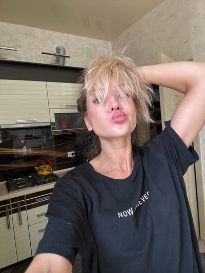 A woman with blonde, messy hair and full lips puckered towards the camera is taking a selfie in what appears to be a kitchen. She is wearing a black t-shirt with the words "NOW NEVER" printed on it in white. The kitchen has light-colored cabinets, a dark countertop with a stove and a yellow kettle with a cherry pattern. The background also shows a textured wall and a doorway. The image is well-lit and appears to be taken during the day. The location is not identifiable as Armavir, Russia, from the image.