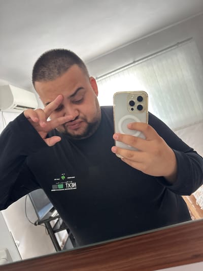A man is taking a mirror selfie in a room that appears to be located in Byala Reka, Bulgaria. He is wearing a black long-sleeved shirt with white text and graphics on it. He has a buzz cut and is making a peace sign with his right hand. He is holding a white iPhone with a clear case in his left hand, which is positioned to capture the reflection. The room has a white wall, an air conditioning unit on the left, and a window with blinds on the right. There is a dark-colored table with a television on it reflected in the mirror. The foreground of the image shows the wooden frame of the mirror.