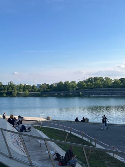 The image shows a view of the Donaukanal in Wien, Austria. It is a sunny day with clear blue skies, with a few clouds in the distance. The Donaukanal is a calm body of water, reflecting the blue sky. Along the riverbank, there are several people relaxing and enjoying the outdoors. Some are sitting on a concrete platform, while others are walking by the water’s edge. There are several trees lining the banks, some of which are in full bloom. The trees create a green and lush backdrop for the scene. The picture is taken from a slight elevation, looking down upon the scene. There is a wire fence in the foreground, separating the elevated platform from the area closer to the water.  The image depicts a tranquil and relaxing atmosphere, with the people enjoying the beautiful scenery.