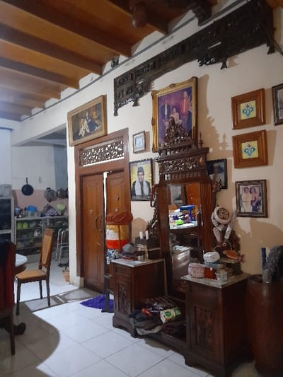 The image shows the interior of a home in Mojokerto, Indonesia, located at the coordinates -7.459 latitude and 112.447 longitude. The room appears to be a living room or bedroom with a large wooden dresser with an ornate mirror. The dresser is filled with various items, including toiletries and decorative items. The room has a white tiled floor. There is a wooden door to the left of the dresser. There are framed pictures on the wall behind the dresser. The walls are painted white and there is a wooden ceiling with beams. There are wooden shelves on the left side of the room and a large wooden table with a chair in the foreground.