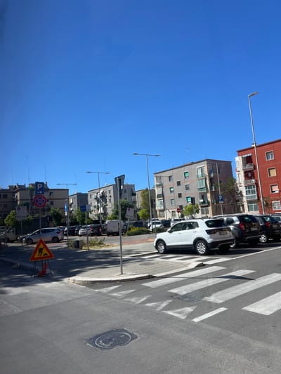 The image, when viewed in its correct upright orientation, depicts a sunlit urban street scene under a clear, bright blue sky. On the right side of the frame, several multi-story apartment buildings with varying facades, balconies, and shutters line the street. Parked cars, including a white SUV and darker vehicles, occupy the street parallel to the sidewalk.
A prominent pedestrian crossing with white zebra stripes spans the asphalt road. Near the crossing, a circular manhole cover is visible. The scene is marked by strong sunlight, casting distinct shadows from buildings, cars, and street furniture onto the ground, indicating a daytime setting, likely mid-day or afternoon.
Numerous street lamps extend across the middle and left portions of the image. Various road signs are visible, including a blue square sign with a white 'P' and an upward arrow indicating parking, a circular blue sign (possibly for mandatory direction), a red circular sign with a diagonal line (no stopping/parking), and a yellow triangular warning sign with a black silhouette of a worker, signaling road work ahead. There are no people visible, and the overall atmosphere is quiet. While specific text on license plates or the manhole cover is not discernible, the road signs are typical of a European urban environment, consistent with the given location context of Bari, Italy.