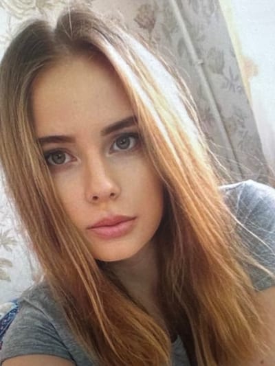 A young woman with long, blonde hair is shown in a close-up, head and shoulders shot. She has fair skin, green eyes, and is wearing a grey t-shirt. The background consists of a patterned curtain. There are no visible indications of the city of Taipei, Taiwan in this image.