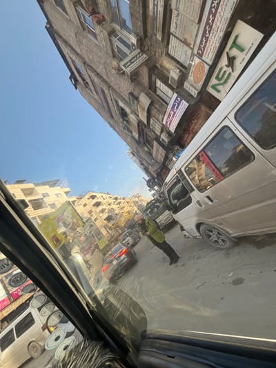 The image shows a street scene with buildings covered in Arabic text signs, including advertisements for "NEXT FOR MOBILE," Dr. Joseph's skin diseases clinic, George Jabara lands, and Dani Finianos. A white van and a person on a motorcycle are on the street. The perspective is from inside a vehicle, with a partially visible dashboard covered with small round stickers or labels.