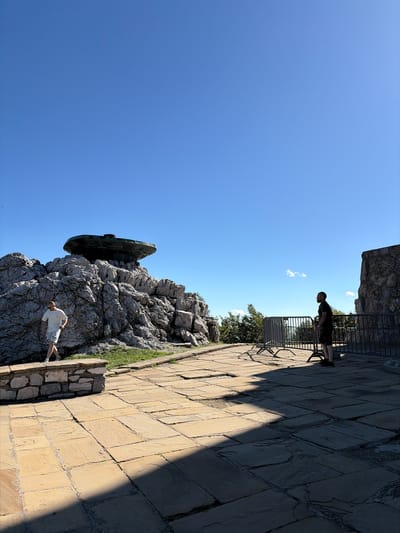 The image depicts a scenic overlook with a view of mountains and a clear blue sky. In the foreground, a paved stone patio is visible, with two men standing at a distance from each other. One man, wearing a white t-shirt and shorts, is walking on the patio. The other man, dressed in a dark t-shirt and shorts, is standing near a metal barrier. Behind the patio, a rocky outcrop with a large, dark, circular structure is prominent. This structure is a monument, likely the Buzludzha Monument, a former communist party headquarters. The surrounding environment includes patches of green grass and trees. The overall impression is one of historical significance and natural beauty.
