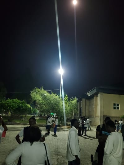 The image depicts an outdoor scene at night, illuminated by a bright street light. A group of people are gathered on a paved walkway in front of a building. The building appears to be a single-story structure with a dark roof and light-colored walls. It has at least two windows visible. To the left of the building are trees and greenery. Some individuals are wearing white clothing, and one person has a black backpack with text on it. The overall impression is of a casual gathering or event taking place in a semi-urban or campus environment. The location is identified as Kason, Nigeria.