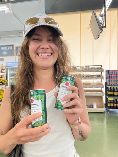 A young woman with long, wavy brown hair and a septum piercing smiles broadly while holding two cans of Tanqueray London Dry Gin & Tonic. She is wearing a white tank top, a light blue baseball cap, and sunglasses. The background appears to be a store, with shelves of products visible. The floor is a light green color. The scene is well-lit, with artificial lighting overhead and natural light filtering in from the side. The location is identified as Pizarra, Spain.