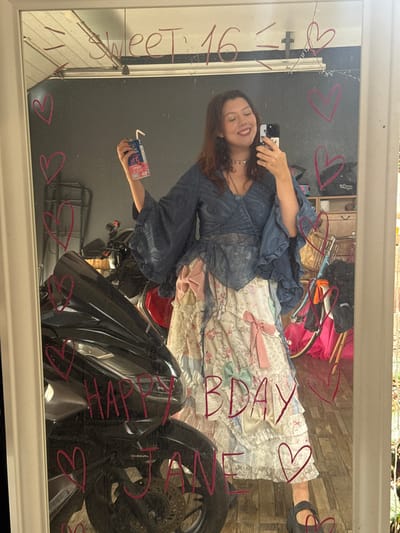 A young woman is taking a mirror selfie in what appears to be a garage or workshop. She is wearing a layered, ruffled skirt and a blue, ruffled top with wide sleeves. In her right hand, she holds a small carton of juice. She is smiling and looking at her phone, which she is holding up to take the picture. The mirror is decorated with pink marker drawings of hearts and the words "Sweet 16" and "Happy Bday Jane". A motorcycle is visible in the reflection of the mirror behind her, along with shelves filled with various items and a bicycle. The floor appears to be wooden planks.