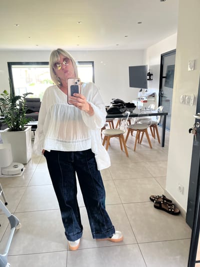 An indoor, full-body mirror selfie captures a woman in a residence located in Saint-Just-Saint-Rambert, France. The woman, with blonde and grey hair and wearing round, light-framed sunglasses, is pouting her lips while holding a light pink smartphone with a dark grip accessory to take the photograph. She is dressed in a loose-fitting, semi-sheer white V-neck blouse, which reveals a dark bra, and dark blue wide-leg jeans with cuffed ankles. Her footwear consists of white shoes with light brown soles. A silver ring is visible on her left ring finger, and her left hand is in her jeans pocket.

The room features light grey tiled flooring, white walls, and a white ceiling with multiple recessed spotlights. In the background on the left, a large window offers a view of outdoor greenery and structures. A large potted plant in a white square planter and a white robotic vacuum cleaner are positioned on the floor nearby. To the right within the reflection, a black rectangular dining table is surrounded by transparent chairs with wooden legs and light-colored seat cushions. A wall-mounted television is visible above a white console table holding various items. On the far right wall, white light switches are mounted, and a set of keys with a red keychain hangs from a door handle. A pair of dark sandals adorned with gold studs rests on the floor adjacent to the wall.