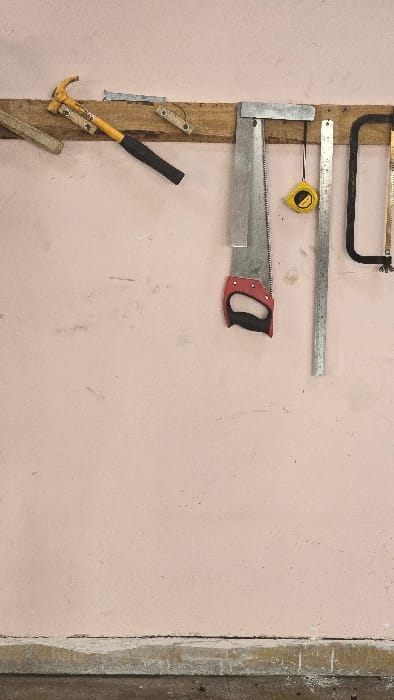 This media file presents a close-up view of several hand tools meticulously organized and hanging on a weathered wooden plank, which is affixed to a pale pink wall.
The subjects are various construction or carpentry tools. From the top, a hacksaw with a black frame is visible. Below it, a long, metallic straightedge or ruler extends horizontally. Directly beneath the ruler, a yellow and black tape measure hangs by its clip, and the brand "STANLEY" is faintly visible on its casing. Further down, a large hand saw with a silver blade and a distinct red and black handle is prominently displayed. Towards the bottom left, a claw hammer with a yellow head and a black handle is positioned diagonally.
The setting appears to be a utilitarian space such as a workshop, garage, or a construction site in Gangapur, India. The wooden plank shows signs of age and use, and the pale pink wall has minor smudges and imperfections, indicating an active working environment. On the far right, a vertical dark brown or grey strip suggests a corner or an adjacent structural element.
No specific activity or event is taking place; the image primarily showcases the organized storage of these tools. The tools themselves appear functional and well-maintained despite visible signs of use. The lighting is even, suggesting an indoor setting or a shaded outdoor area, and no specific time of day or weather conditions can be discerned. There are no people or explicit emotions conveyed.