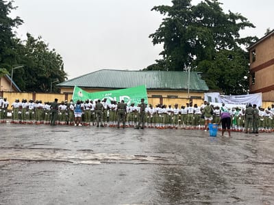 A group of people, many wearing matching green uniforms with white shirts and hats, stand in rows outdoors. They appear to be part of an organized event, possibly a ceremony or parade. The ground is wet and appears to be a paved surface. In the background, there are buildings with green roofs and a large tree. A banner with text is visible, and the sky is overcast, suggesting a cloudy or rainy day. The overall mood seems neutral, focused on the activity at hand. Some individuals are wearing military-style uniforms, while others are in what looks like civilian attire. One person is seen interacting with a blue bin. The location is identified as Alagba, Nigeria. The visible text on the banner includes "I AM PROUD TO BE A" and "NYSC KUDIMATA MARKET".