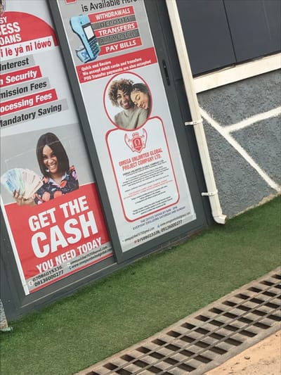 The image displays two vertical advertising banners affixed to the exterior of a commercial building in Akure, Nigeria. The banners promote "OMEGA UNLIMITED GLOBAL PROJECT COMPANY LTD." The left banner, predominantly red and white, features a smiling woman holding a fan of Nigerian Naira banknotes with the prominent text "GET THE CASH YOU NEED TODAY." Partial text mentions "ESS LOANS," "Interest," "Security," "Mission Fees," "Processing Fees," and "Mandatory Saving." The right banner, white with red accents, lists services such as "WITHDRAWALS, DEPOSITS, TRANSFERS, AIRTIME REFILLS, PAY BILLS," accompanied by an image of a mobile POS terminal. This banner also includes a circular photograph of two smiling women and the company logo. Text below indicates "Quick and Secure," accepting "debit cards and transfers," and "POS Transfer payments." The company is identified as "OMEGA UNLIMITED GLOBAL PROJECT COMPANY LTD." based in "Akure, Ondo State, Nigeria." Office hours are stated as "9AM - 5PM EVERYDAY EXCLUDING SATURDAY AND SUNDAY." Both banners provide contact phone numbers (07086015336, 09136000277, U/0806015336), an email (omega.global7070@gmail.com), and a website (www.omegaunlimitedglobal.com). The building's exterior features dark gray paneling and a white vertical downspout. Below the banners, a strip of artificial green turf is visible, adjacent to a ground-level metal drainage grate in the foreground.