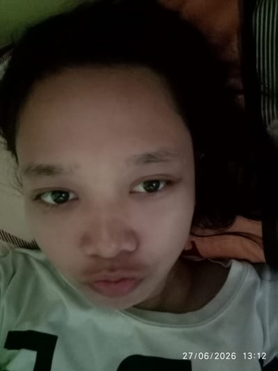 A close-up, selfie-style photograph shows a young person's face filling most of the frame. The subject has dark hair that partially covers their forehead and sides of their face. Their eyes are open, and their mouth is slightly pursed. They are wearing a light-colored t-shirt with a dark graphic design on the left side of the chest. The background appears to be a bed with a striped pillow and a green blanket. The lighting is dim, suggesting the photo was taken indoors. A timestamp in the bottom right corner reads "27/06/2026 13:12". The image does not contain any identifiable landmarks or features that would indicate a specific location within Sukaraja, Indonesia.