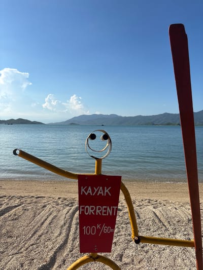An outdoor shot captures a whimsical, homemade signpost on a beach in Vĩnh Lương, Vietnam. The signpost, fashioned to resemble a stick figure, is bright yellow with a red rectangular sign attached to its torso. The sign reads "KAYAK FOR RENT 100K/60m" in white lettering. The stick figure has a round, metal face with two large eyes and a smiling mouth. Its arms are outstretched, with one arm ending in a circular opening. To the right of the stick figure, a large, red kayak paddle stands upright, partially obscuring the background.
The beach itself is covered in a mixture of sand and small pebbles, with distinct patterns of light and shadow suggesting the late afternoon sun. Beyond the beach, the calm, blue ocean stretches out, meeting a line of verdant, tree-covered hills that form the horizon. The sky is a clear blue, dotted with a few fluffy white clouds. The overall atmosphere is one of a relaxed, seaside destination, likely a tourist spot offering recreational activities.