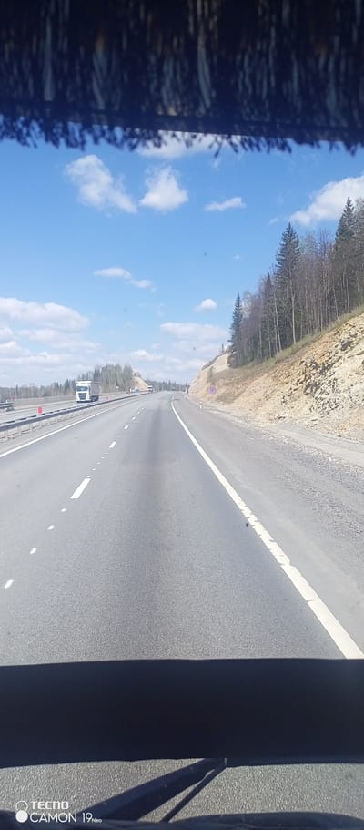 The image depicts a highway in Myasnikovo, Russia, under a bright blue sky with scattered clouds. The view is from inside a vehicle, looking out over the road. The highway has multiple lanes with white painted lines dividing them. A truck is visible in the distance on the left side of the road. To the right of the highway is a hillside with trees. The foreground shows the dashboard of the vehicle, partially obscuring the lower part of the scene. A sun visor fringe hangs from the top of the frame. A "no stopping" sign is visible on the hillside along the right side of the road. The asphalt is dark gray and shows signs of wear.