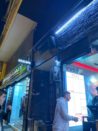 An nighttime, horizontally-oriented image depicts a vibrant street food or bar scene in Koridallos, Greece. The perspective is slightly upwards, capturing the exterior of an establishment illuminated by various artificial lights.
On the right side of the frame, a man wearing a light-colored hoodie stands, looking upwards at a large, brightly lit digital menu board. The board displays a grid pattern with the word "MENU" visible at its top left corner. Below the man's gaze, partially obscured, is a dark counter area. Further down and to the right, another person, with a hat and dark jacket, holds a green bottle, possibly of wine or champagne, their face partially visible.
The establishment features a prominent brick-textured facade with a long fluorescent light fixture running along its length, casting a cool blue glow. Below this, various fixtures are mounted, including a rounded black speaker and a wire mesh cage. Several illuminated signs are visible: a black rectangular sign reads "DEALERS" with "REAL STREET FOOD" written beneath it, and a green neon sign displays "BRAKOU LIAS" with "ATTHENS" below it, confirming the Greek location. The dark sky is visible on the left side of the image, contrasting with the bright lights of the commercial space. The overall impression is one of an active urban night scene.