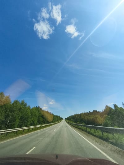 The image depicts a view from inside a vehicle looking forward on a paved road. The road is divided by white lane markings and curves slightly towards the right as it recedes into the distance. A metal guardrail runs along the right side of the road, bordering a dense forest of green trees. On the left side of the road, another guardrail is visible, also separating the road from a similar tree line. The sky above is a vibrant blue with a few scattered white clouds and a bright sun with lens flare in the upper right quadrant. The overall scene suggests a drive through a rural or natural landscape in Russia, possibly near Большая Уса, Russia given the context, during daylight.