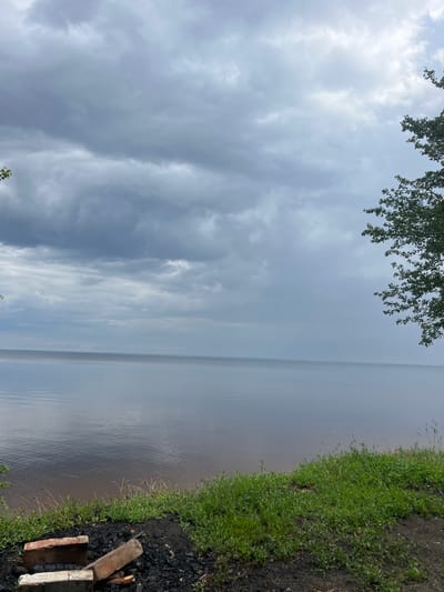 The image depicts a wide, calm body of water under a cloudy sky. The water is a muted gray, reflecting the overcast conditions. The shoreline in the foreground is covered with green grass and some darker soil with scattered bricks and what appears to be remnants of a fire pit. To the right, the branches of a green tree overhang the scene. The horizon line is distinct, separating the water from the sky, which is filled with various shades of gray clouds. The overall impression is a quiet, natural setting. The location is Lyutizh, Ukraine.