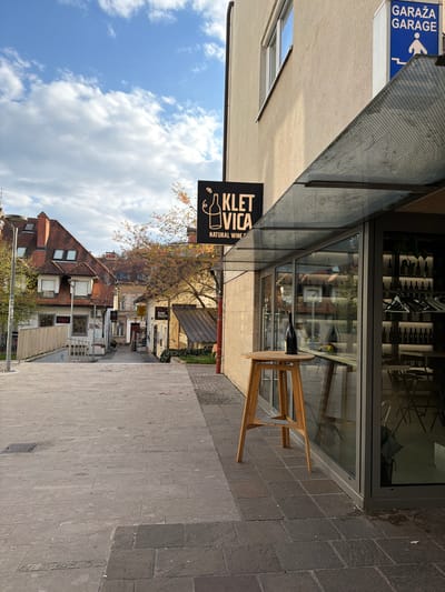 The image depicts an urban street scene in Ljubljana, Slovenia. The perspective is slightly tilted. A building on the left has a sign that reads "GARAŽA GARAGE" and another sign for "KLET VICA NATURAL WINE" hanging below. A glass window displays wine bottles and reflections of the buildings and sky. A small, round wooden table with a bottle of wine on it stands on the sidewalk next to the building. The street extends into the background, lined with buildings and trees. Above, a blue sky with white clouds is visible. The street is paved with gray stone. The overall impression is that of a charming European street with a wine bar.