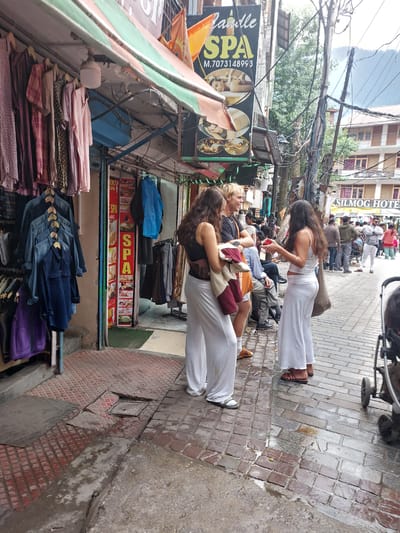 The image shows a street scene in Manali, India, likely in the main market area. In the foreground, three young women are standing on a brick-paved sidewalk. One woman on the left wears a long white skirt and a dark top with a brown bag over her shoulder. The middle woman, a blonde, wears a white skirt and a dark top, holding a garment in her arms. The woman on the right also wears a long white skirt and a white tank top, carrying a large woven bag. They appear to be conversing.
Adjacent to them is a storefront displaying clothing on hangers, including denim shirts and purple garments. Further down the street, a spa with the name "Candle SPA" is visible, featuring images of massage services. Behind the spa, buildings with balconies and signs, including "SILMOG HOTEL," can be seen. The street is populated with various individuals, some walking and others gathered in groups. Power lines and utility poles are prominent overhead. The ground is wet, suggesting it may have recently rained. A stroller is partially visible on the right side of the frame. The overall atmosphere suggests a busy commercial street in a tourist destination.