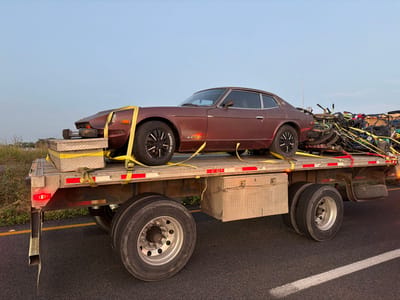 A maroon Datsun 280Z is secured with yellow straps atop a flatbed trailer. Silver diamond-plate toolboxes are strapped to the car's hood. The trailer has dual wheels and silver toolboxes affixed underneath. In the trailer's rear, a jumble of yellow-strapped items, including bikes and ATVs, are visible. The scene unfolds on an asphalt road with greenery along the horizon under a clear sky.