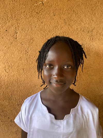 A young girl with dark skin and braided hair stands in front of a textured, ochre-colored wall. She is wearing a white V-neck shirt with short sleeves. The girl is looking directly at the camera with a slight smile. The lighting suggests an outdoor setting, possibly in the city of Riyom, Nigeria, given the style of the wall and the attire.