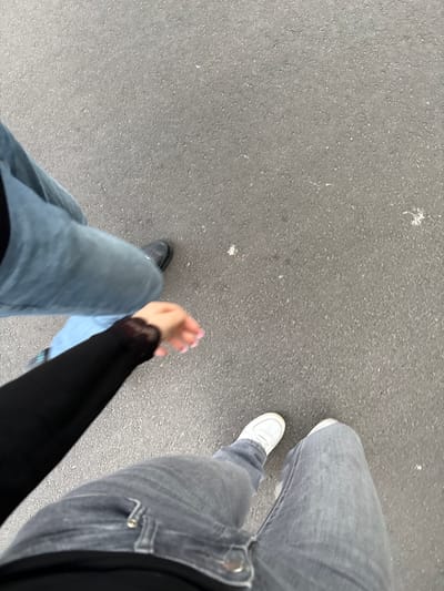 The image shows a point-of-view shot of two individuals walking on an asphalt surface, likely a street or pavement, in Pori, Finland. One person, whose legs are clad in light blue jeans and black shoes, is walking ahead and slightly to the left. The other person, wearing gray jeans and white sneakers, is walking behind and to the right. They are holding hands; one hand, with painted nails, is visible on the right side of the frame, adorned with a black lace cuff. The environment is an outdoor urban setting, with the asphalt providing the primary visual element, suggesting a walkway within the city.