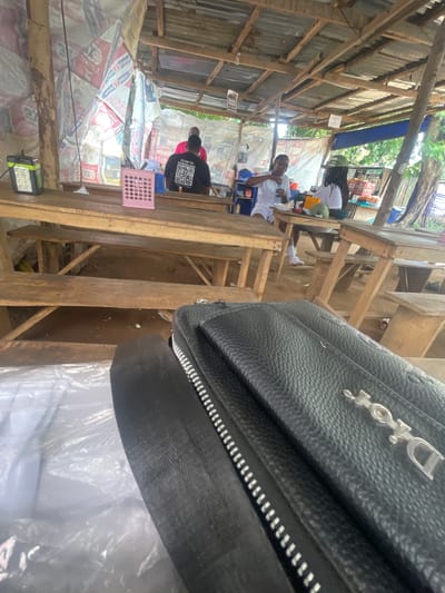 The image depicts an outdoor dining area, likely at a roadside establishment in Orun Ekiti, Nigeria. Several wooden tables and benches are arranged under a rustic, open-air shelter constructed with corrugated metal roofing and wooden beams. The walls of the shelter are draped with what appear to be tarpaulins or repurposed materials, some bearing text and graphics.

In the background, three individuals are visible. One person, wearing a black t-shirt with a QR code and the text "SCAN TO DOWNLOAD," is seated at a table. Another person, in a white shirt, is gesturing with their left hand while seated at a table. A third individual, also in a white shirt, is seated further back.

On the tables, various items are present, including cups, plates, a pink basket containing utensils, and bottles. To the left of the frame, a solar-powered lamp with multiple LED panels is placed on a table.

In the foreground, a black leather bag with a prominent "Dior" logo is placed on top of another object, partially obscuring the view of the dining area. The overall environment suggests a casual, possibly humid climate.