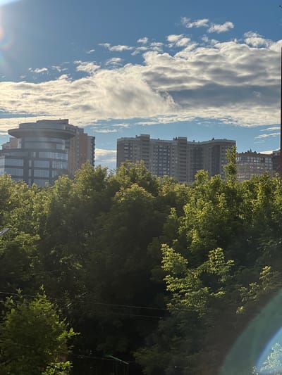 This media file captures a cityscape with a focus on residential buildings and lush green trees under a bright, cloudy sky. The perspective is vertical, suggesting the image was taken from a high vantage point or that the phone was held upright.

The scene is dominated by a large, dense tree on the right, its leaves illuminated by sunlight, creating a vibrant green contrast against the blue sky. Behind the tree, a series of multi-story residential buildings are visible. These buildings vary in architectural style, with some appearing to be older, block-like structures, and others featuring more modern designs with glass facades. The buildings stretch across the frame, indicating a densely populated urban area.

The sky is filled with dynamic, elongated clouds that stretch horizontally, with smaller, scattered clouds interspersed. The sunlight filtering through the clouds suggests it is daytime, likely morning or afternoon given the brightness. There are no visible people in the image.

Notable details include a slight lens flare or light streak in the upper right corner, indicating the direction of the sun. A subtle rainbow-like effect is visible in the lower left corner, possibly a result of the light interacting with the camera lens. The presence of power lines in the lower half of the frame suggests an urban or suburban setting. No text is discernible in the image. The overall impression is one of urban life integrated with abundant greenery, under a dramatic sky. The location context of Pushkino, Russia, aligns with the visual cues of urban apartment blocks and trees.