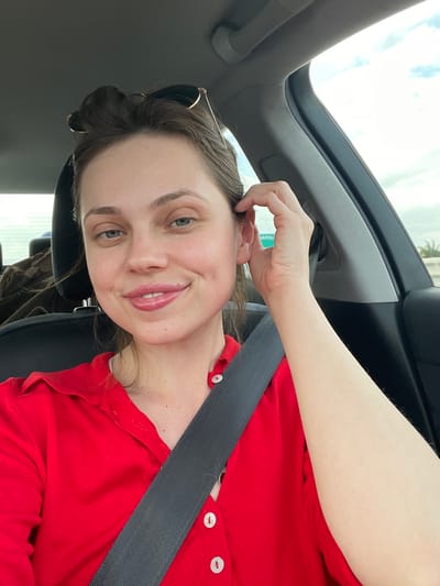 The image displays an adult female situated in the passenger seat of a vehicle, located in Scafati, Italy. The female subject has light skin, brown hair styled in an updo, and brown eyes, with sunglasses perched on her head. She is wearing a red collared shirt with white buttons and is secured by a dark-colored seatbelt extending diagonally across her chest. Her gaze is directed towards the camera, and she exhibits a slight, closed-mouth smile. Her right hand is raised, with fingers lightly touching the area near her right ear. Through the vehicle's right-side window, a partial green road sign is visible, displaying "NAPOLI" in white lettering, indicating a route towards Naples. The background outside the vehicle shows a cloudy sky and distant vegetation. Inside the car, behind the driver's seat, an object covered with dark fabric is partially visible.