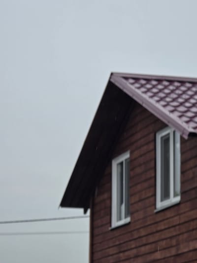 A blurred, medium-angle shot captures the upper section of a wooden house with a red, shingled roof under an overcast sky. The house has horizontal brown wood siding and two white-framed windows visible on the upper floor. A thin, dark wire stretches horizontally across the left side of the frame, suggesting its location in a populated area, possibly the city of Ермолаево, Russia. There are no people or animals visible.