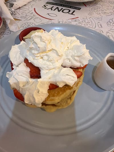 A high-angle, close-up photograph depicts a stack of three golden-brown pancakes presented on a round, light blue ceramic plate. The pancakes are heavily topped with a substantial quantity of white whipped cream, interspersed with bright red slices of strawberries. To the right of the pancake stack, a small, white ceramic pitcher contains a dark amber liquid, presumed to be syrup. To the left, a red ramekin containing a pale yellow substance is partially visible. The plate rests on a white paper placemat patterned with various black line drawings of food items, including pizza and cheese. The placemat also displays an inverted red semi-circular logo with black text, partially visible, which reads "TONY'S COFFEE SHOP & RESTAURANT." This scene is located at Tony's Coffee Shop & Restaurant in Tirana, Albania. No individuals or active interactions are visible.
