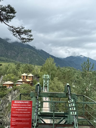 The image depicts a suspension bridge in the city of Chemal, Russia, leading to Patmos Island. The bridge structure is painted green and is made of metal with thick cables supporting the walkway. The walkway itself appears to be made of wooden planks and is partially visible. In the foreground, a red sign with white lettering displays a warning in Russian for visitors to Patmos Island, cautioning them about the area and listing rules. Several people are visible on the bridge, and more are gathered near some wooden buildings on the rocky island. The background features lush green forests and majestic mountains under a cloudy sky. The overall scene suggests a tourist destination with a blend of natural beauty and man-made infrastructure.