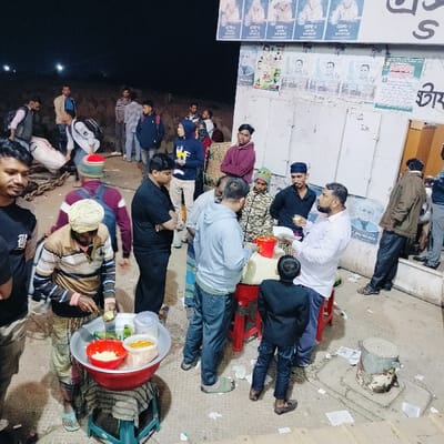This image captures a nighttime street scene in Bhola, Bangladesh, depicting a bustling outdoor food stall environment. Approximately 15 individuals, predominantly adult males, are present.
In the foreground, to the left, a male vendor wearing a striped shirt and a lungi is actively preparing food. He uses a knife to slice a green vegetable, likely a cucumber, into a red bowl which contains other white and red ingredients. This large red bowl rests on a metallic stand, which in turn sits on a blue plastic stool. Adjacent to this, on a red plastic stool, is another food setup with a man in a light grey hoodie looking down at a red bucket. An adult male in a white shirt holds a small white dish, appearing to consume food. A young boy in a dark jacket stands facing this food preparation area.
The background features a light-colored building wall densely covered with numerous posters featuring Bengali text and portraits of men, suggesting political or social advertisements. Further to the left, multiple individuals are standing or walking on what appears to be a slightly elevated, rocky or earthen embankment. Heavy metal chains are visible on the ground in the far left, suggesting proximity to a dock or mooring area. The ground surface is concrete or paved and shows scattered debris, including discarded papers. A rusted metal bollard is visible on the right side of the frame. Artificial lighting illuminates the entire scene under a dark night sky.