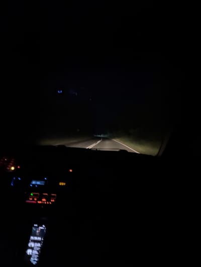 The image depicts a night scene, likely from inside a vehicle moving on a road near Cherni Vrah, Bulgaria. A dimly lit road is visible through the windshield, with a grassy area to one side. The interior of the vehicle is visible in the foreground, including the dashboard with illuminated displays and controls. No people are clearly visible. The environment appears to be rural or sparsely populated, given the darkness and lack of prominent urban features. The main action inferred is the movement of the vehicle on the road at night.