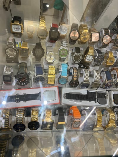 This close-up, high-angle shot captures a crowded retail display case filled with numerous watches, likely within a market or store setting in Lusaka, Zambia. The watches are presented in various styles, sizes, and materials, suggesting a wide selection for sale.

The display case is illuminated by bright overhead lighting, creating significant glare and reflections on the glass surface, which partially obscure some of the items and the background. Watches are arranged densely, often overlapping or displayed on stands, with some still in their original packaging or boxes.

The collection includes a diverse range of timepieces:
*   **Analog watches:** featuring round, square, and rectangular faces with various dial designs, including chronograph styles and simple hour markers.
*   **Digital watches:** one prominent Casio digital watch is visible near the bottom center.
*   **Band materials:** a mix of metal link bracelets (gold-toned, silver-toned, and two-tone), leather or synthetic straps (black, brown, blue), and silicone bands (orange, light blue, red).
*   **Colors:** predominant colors are gold, silver, black, and rose gold for cases and bands, with some faces featuring vibrant hues like green, blue, and white.

Notable items include an unpacked black watch strap and case (reminiscent of an Apple Watch accessory) and an orange-banded watch, both in rectangular white packaging. Small price tags or labels are sporadically visible beneath some watches, but the text on most is unreadable.

Visible text and brand names include "TOMI" on a watch near the top left, "RENA..." on a box near the top center, and "CASIO" on a digital watch at the bottom center. A small tag with "GUESS" is also visible near the bottom right center. The overall impression is one of abundance and variety, characteristic of a bustling market stall or a casual watch vendor.