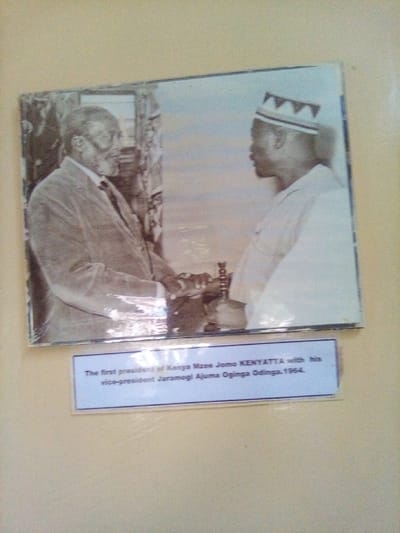 A historical photograph, laminated and showing signs of age or toning, is mounted on a light-colored wall alongside a blue descriptive caption. The photograph depicts two African men. On the left, an older man with a beard and glasses, identified as Mzee Jomo Kenyatta, wears a light-colored suit jacket and tie, facing towards the right. On the right, a younger man, identified as Jaramogi Ajuma Oginga Odinga, wears a light-colored tunic and a kofia with a zigzag pattern on its trim, facing towards the left. Both men have their hands extended towards the center, appearing to engage in a handshake or exchange a small, dark cylindrical object. The background within the photograph is indistinct, with darker, textured elements on the left and a lighter, more open area on the right. The blue caption below the photograph reads: "The first president of Kenya Mzee Jomo KENYATTA with his vice-president Jaramogi Ajuma Oginga Odinga. 1964." This entire display is physically located in South Sakwa ward, Kenya.