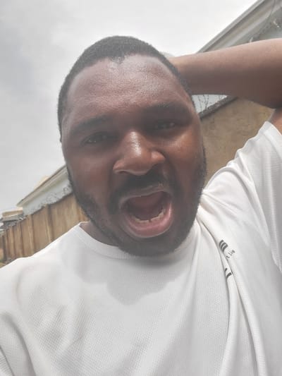 A close-up, rotated image shows a dark-skinned Black man with a beard and short hair, wearing a white t-shirt with a logo that reads "Smart Life". The man has his mouth open wide. His right arm is raised and resting on the back of his head. In the blurred background, there is an outdoor stairwell on a building with a razor wire visible. The location is Suleja, Nigeria.