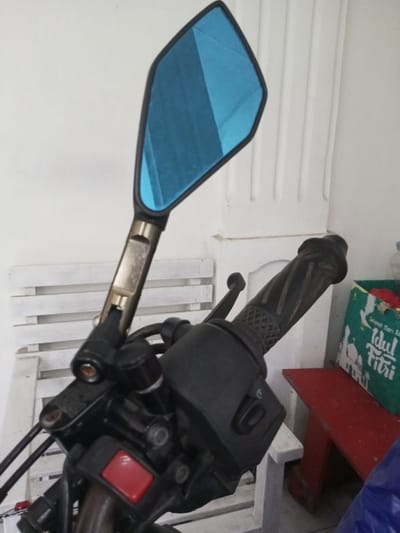 A close-up shot focuses on the handlebar of a motorcycle. The left side features a blue-tinted mirror with a black frame, connected to the handlebar by a silver arm and black joint. The motorcycle's controls are visible, including a red kill switch and black plastic housing with buttons. The black rubber grip is textured. In the background, a white slatted bench and the corner of a red table are visible, along with a green bag with an Eid al-Fitr graphic.